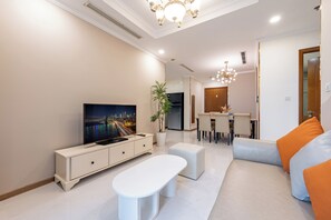 A modern living room with a flat-screen TV, a dining area, and a seating area with a sofa and ottoman. - Vinhomes Cozy Apartment (Ho Chi Minh City)