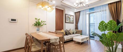 A modern living room with a wooden dining table, sofa, and large windows with curtains.