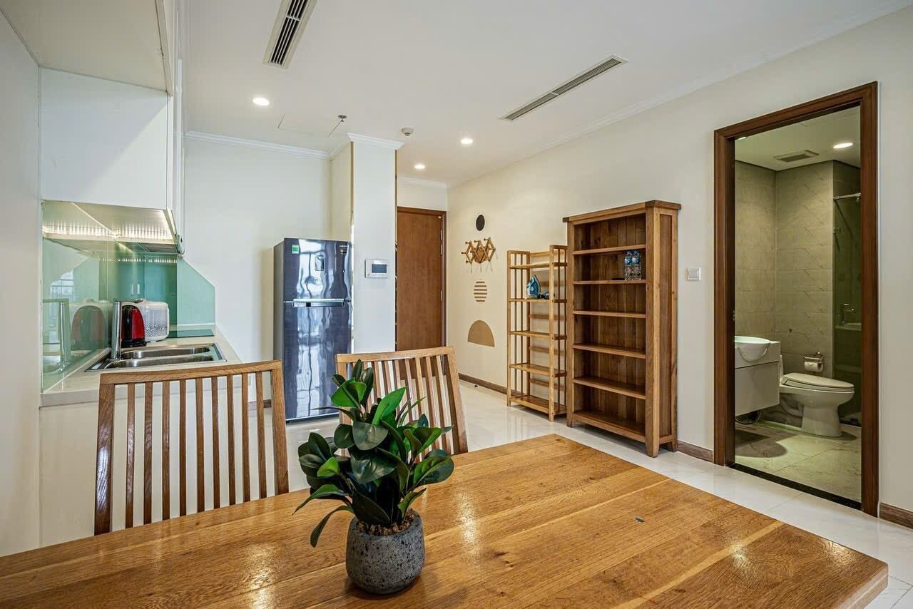 Eine moderne Küche mit Kühlschrank, Mikrowelle und Spüle. Ein Esstisch mit Stühlen und ein Blumentopf sind vorhanden. Eine Holzregal steht an der Wand, und durch eine offene Tür ist ein Badezimmer zu sehen.