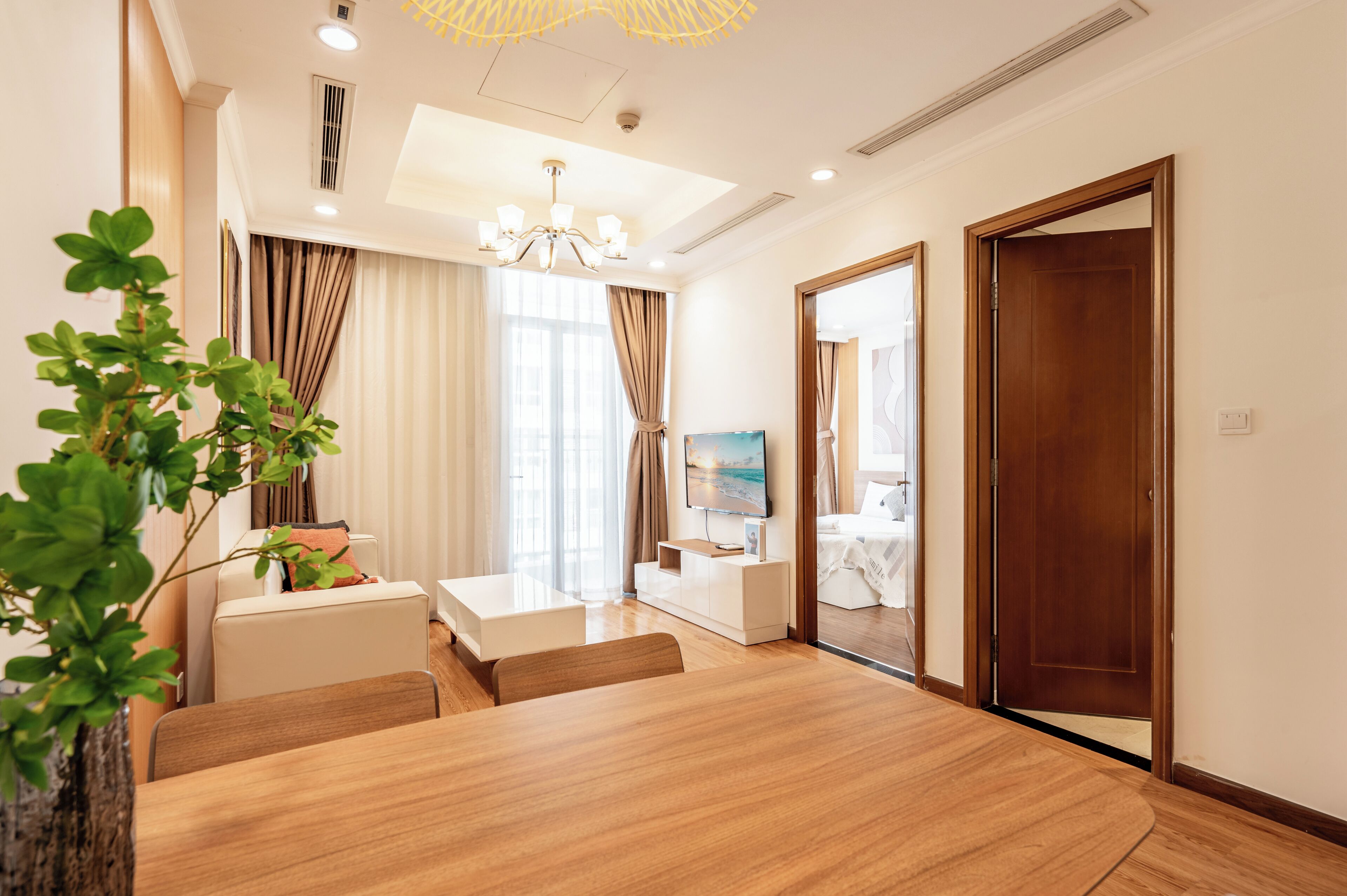 Una habitación de hotel moderna con una mesa de comedor de madera, un sofá, un televisor y un ventanal con cortinas.