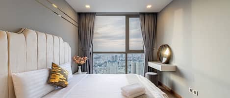 Un hotel moderno con una cama grande, vistas a la ciudad, un tocador con espejo y una pequeña mesa con un jarrón de flores.