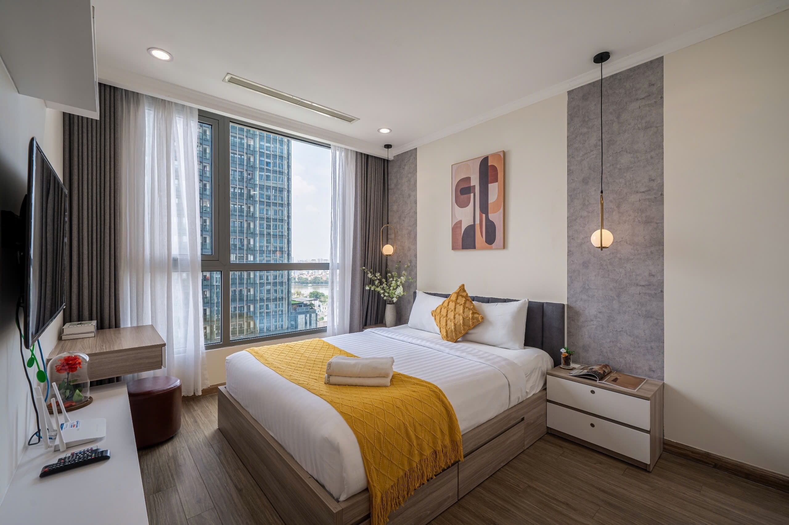 A modern hotel room with a large window, a bed with a yellow throw, a bedside table, a nightstand, and a flat-screen TV.