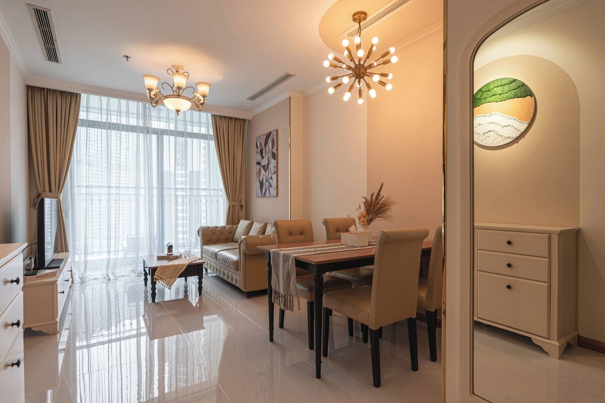 A well-lit living room with a sofa, dining table, and chairs, a chandelier, and a painting on the wall.