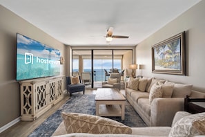 A spacious living room with a large TV, comfortable seating, a ceiling fan, and a view to the outside.