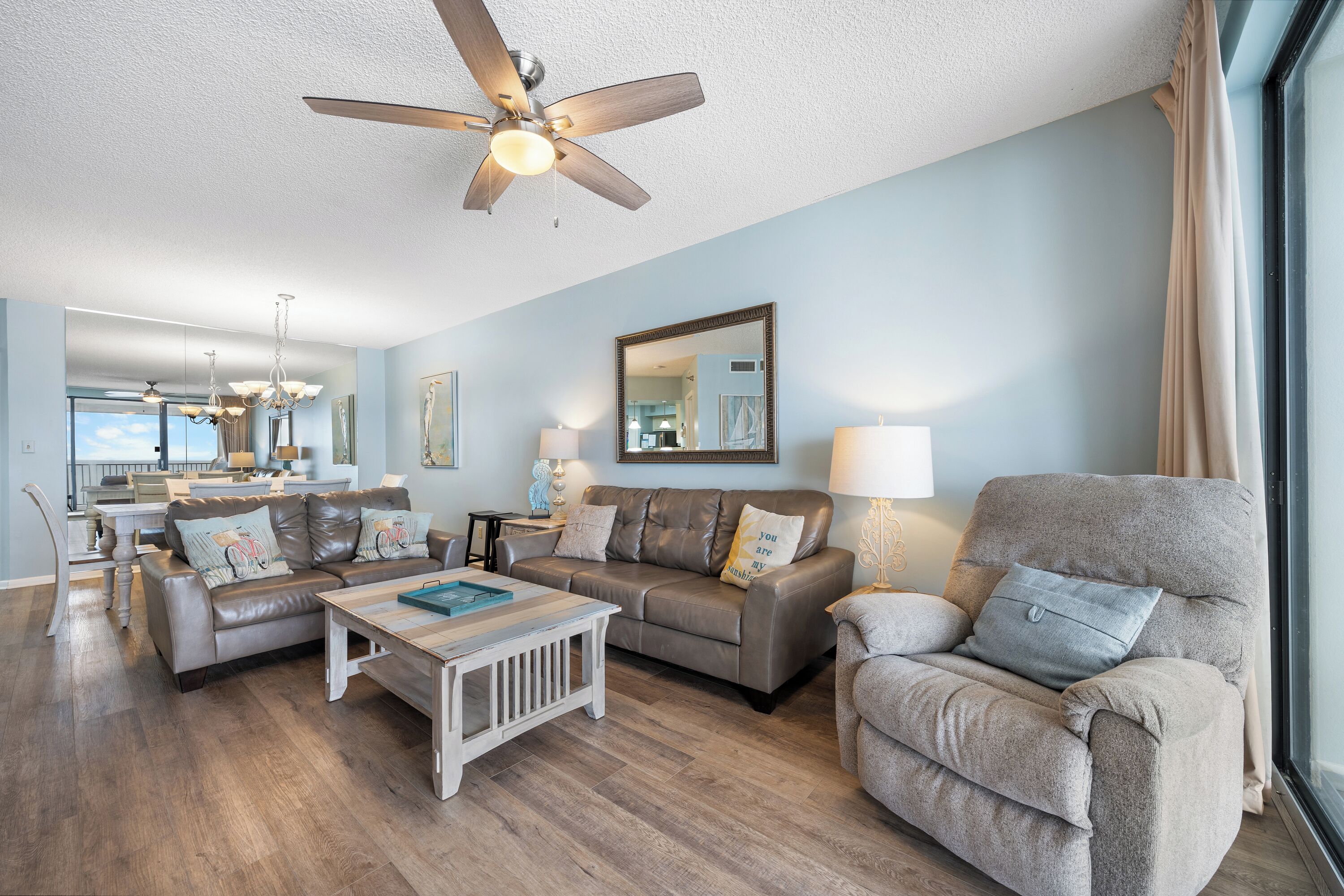 A spacious living room with a ceiling fan, a dining area with a kitchen in the background, and a large window with curtains.
