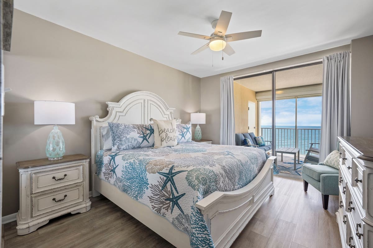 Une chambre à coucher comprenant un lit, des tables de chevet, une commode, un ventilateur de plafond et un balcon avec vue sur l’océan.