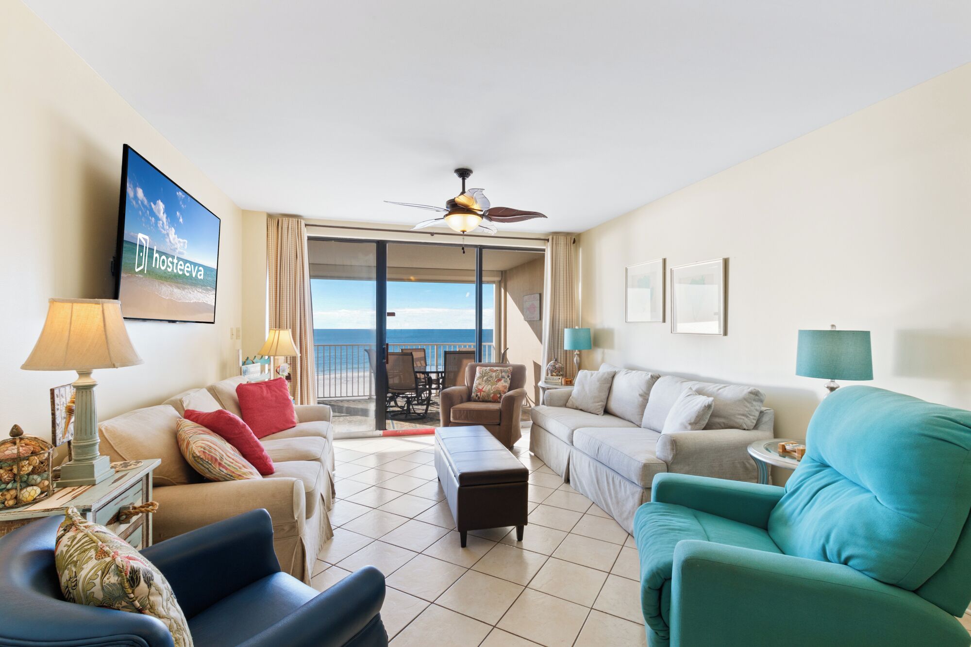 A spacious living room with a large sofa, a smaller armchair, a coffee table, and a television mounted on the wall. There is a balcony with a view of the ocean.