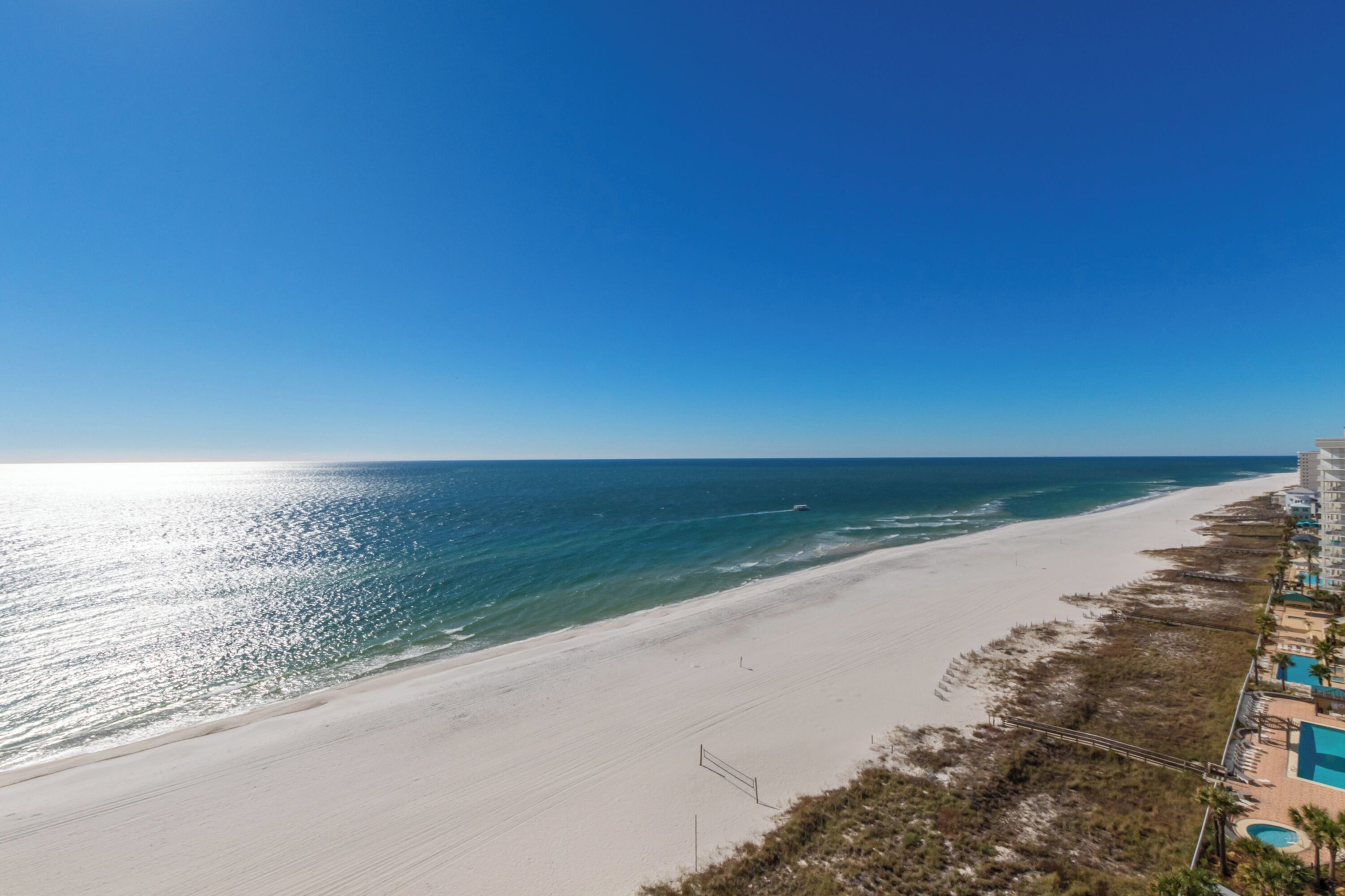 A beach view with a clear sky, a sandy beach, and a building with a pool on the right.