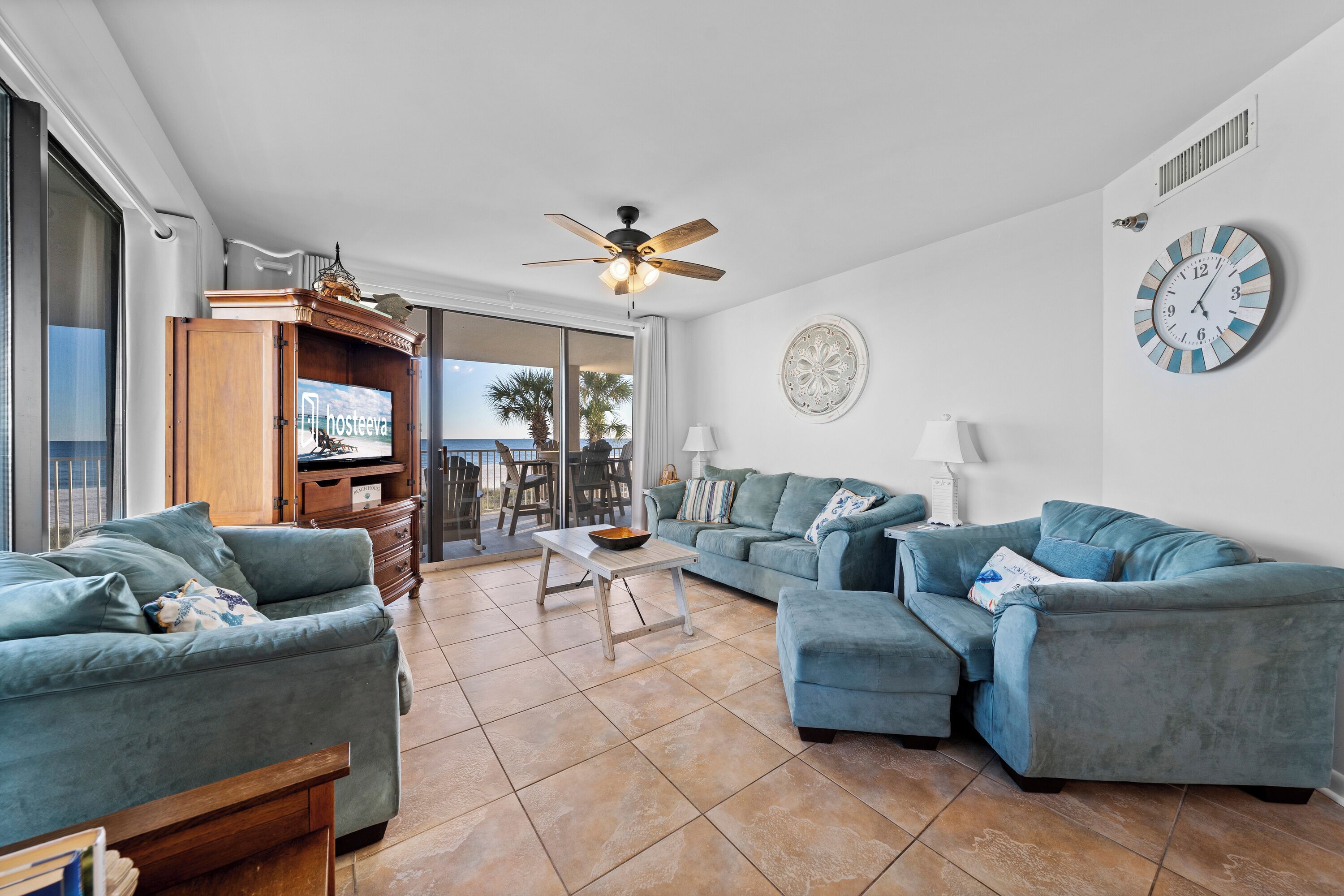 A spacious living room with a ceiling fan, a large round clock, and a view of the ocean through the sliding glass doors.