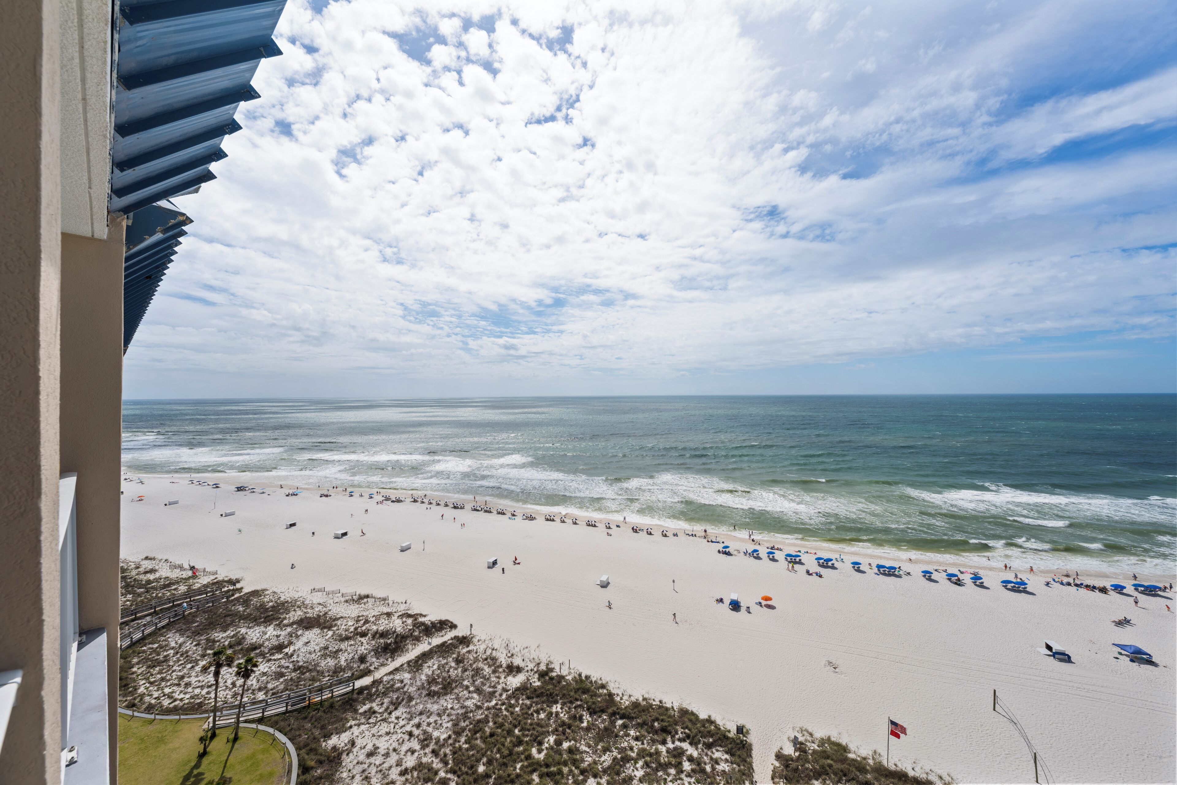 A beach view from a balcony with a railing, ocean, and beachgoers.