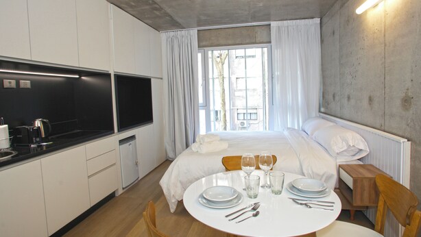 A modern hotel room with a kitchen area, a dining table set for two, and a large bed with white linens. - Urban Rent (Buenos Aires)