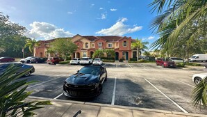 A parking lot with several cars, a building with a red facade, and palm trees.