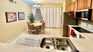 A kitchen with a stainless steel sink, a microwave, a refrigerator, wooden cabinets, and a dining area with a table and chairs.
