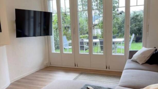Een ruime woonkamer met een groot raam, een flatscreen televisie aan de muur en een bank met een afstandsbediening erop.