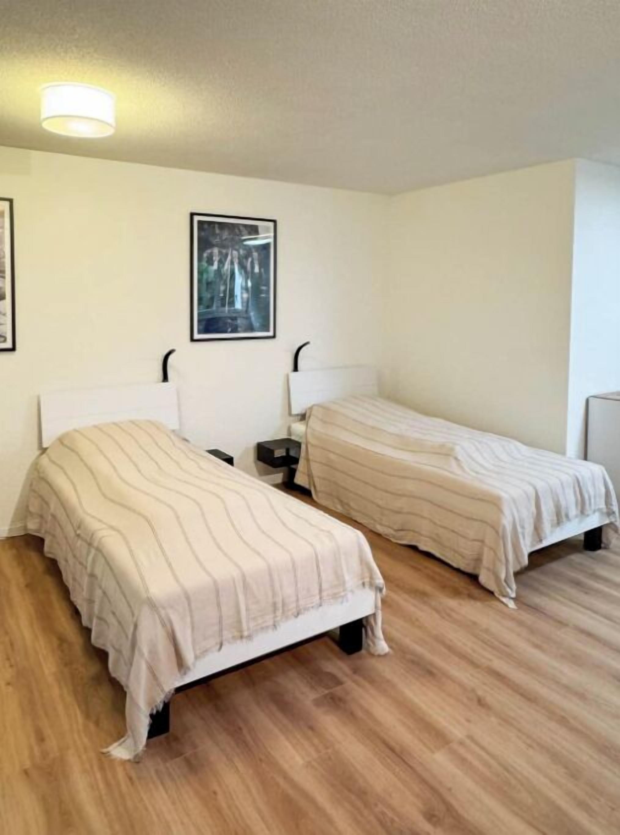 Une chambre avec deux lits, chacun doté d’une tête de lit et d’une table de chevet. La chambre est pourvue d’un plancher en bois et d’un luminaire au plafond.