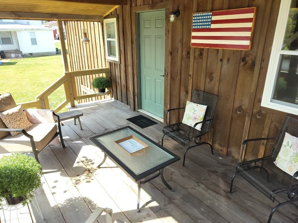 Une véranda en bois avec une table et des chaises, une porte verte et un drapeau américain.