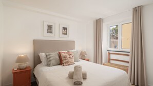 Un lit bien fait, avec une couette blanche et des serviettes pliées, une tête de lit en bois, deux tables de chevet avec des lampes et une fenêtre avec des rideaux.