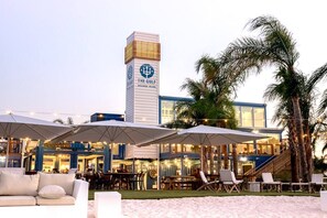 Un hotel frente al mar con terraza al aire libre, palmeras y un edificio alto con un logotipo.