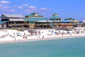 Un hotel frente a la playa con varias edificaciones de techos verdes, una playa de arena y un cielo azul despejado.