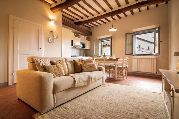 A cozy living room with a sofa, a dining table, and chairs. There is a kitchen area in the background, a window with a view, and a radiator under the window. - RIDOLFO HAUS - Apartment in San Miniato (San Miniato)