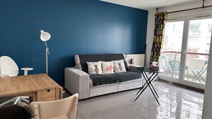 Ein modernes Wohnzimmer mit blauer Wand, einem grauen Sofa mit dekorativen Kissen, einem Holzesstisch, einem kleinen runden Tisch und einem Balkon mit Aussicht.