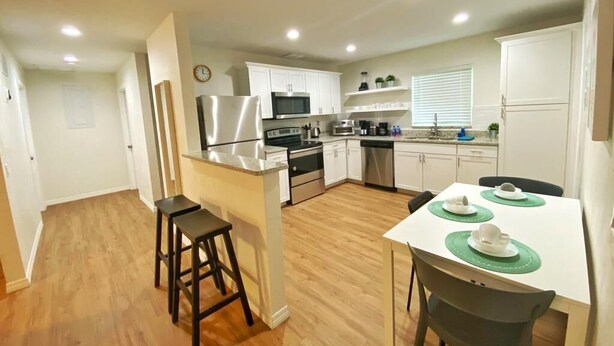 Una cocina moderna con armarios blancos, electrodomésticos de acero inoxidable y un comedor con mesa blanca y taburetes de bar.