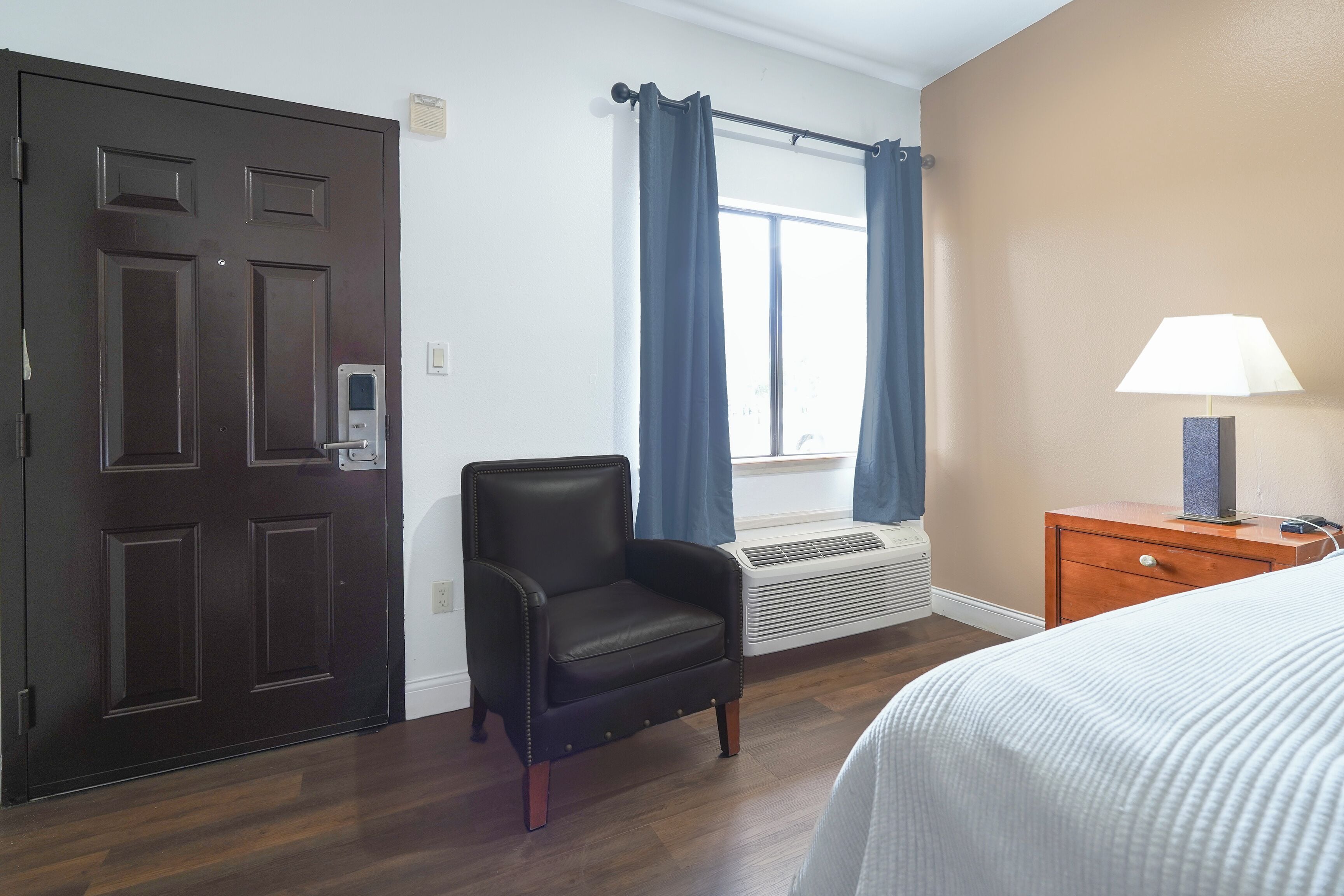 Une chambre d’hôtel avec une porte en bois foncé, un fauteuil en cuir, un lit recouvert d’une courtepointe blanche, une table de chevet en bois avec une lampe, et une fenêtre ornée de rideaux bleus.