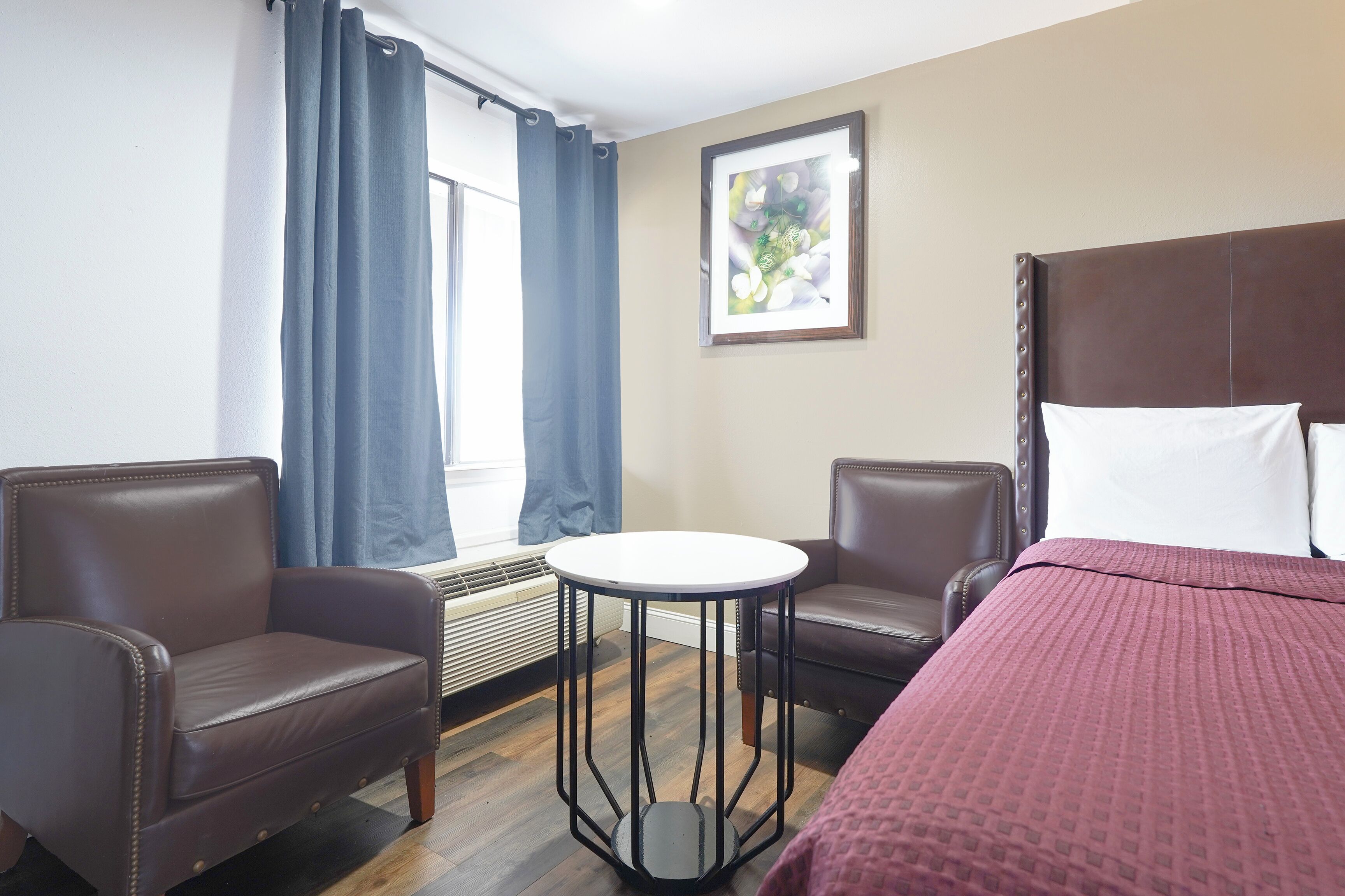 A hotel room with a bed, two armchairs, a small round table, a window with curtains, and a framed picture on the wall.