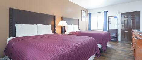 Habitación de hotel con dos camas, suelo de madera, mesita de noche con lámpara y puerta con espejo.
