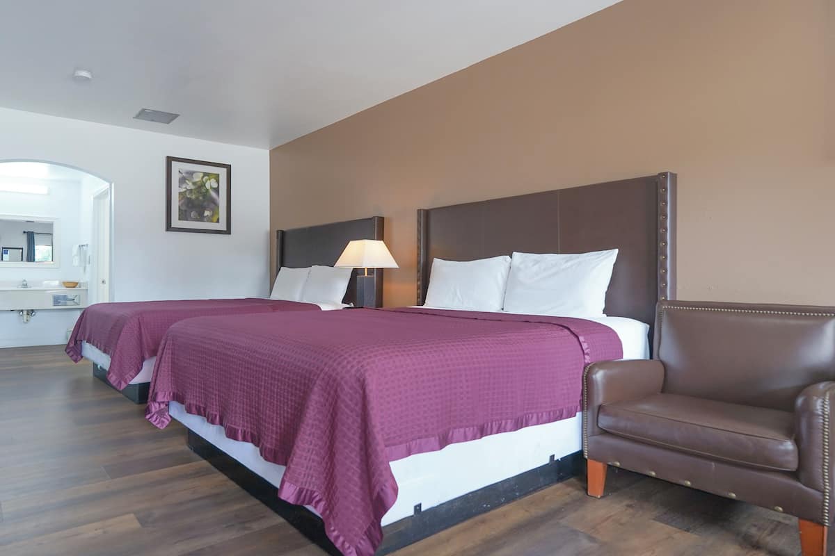 A hotel room with two beds, a brown leather armchair, and a nightstand with a lamp.
