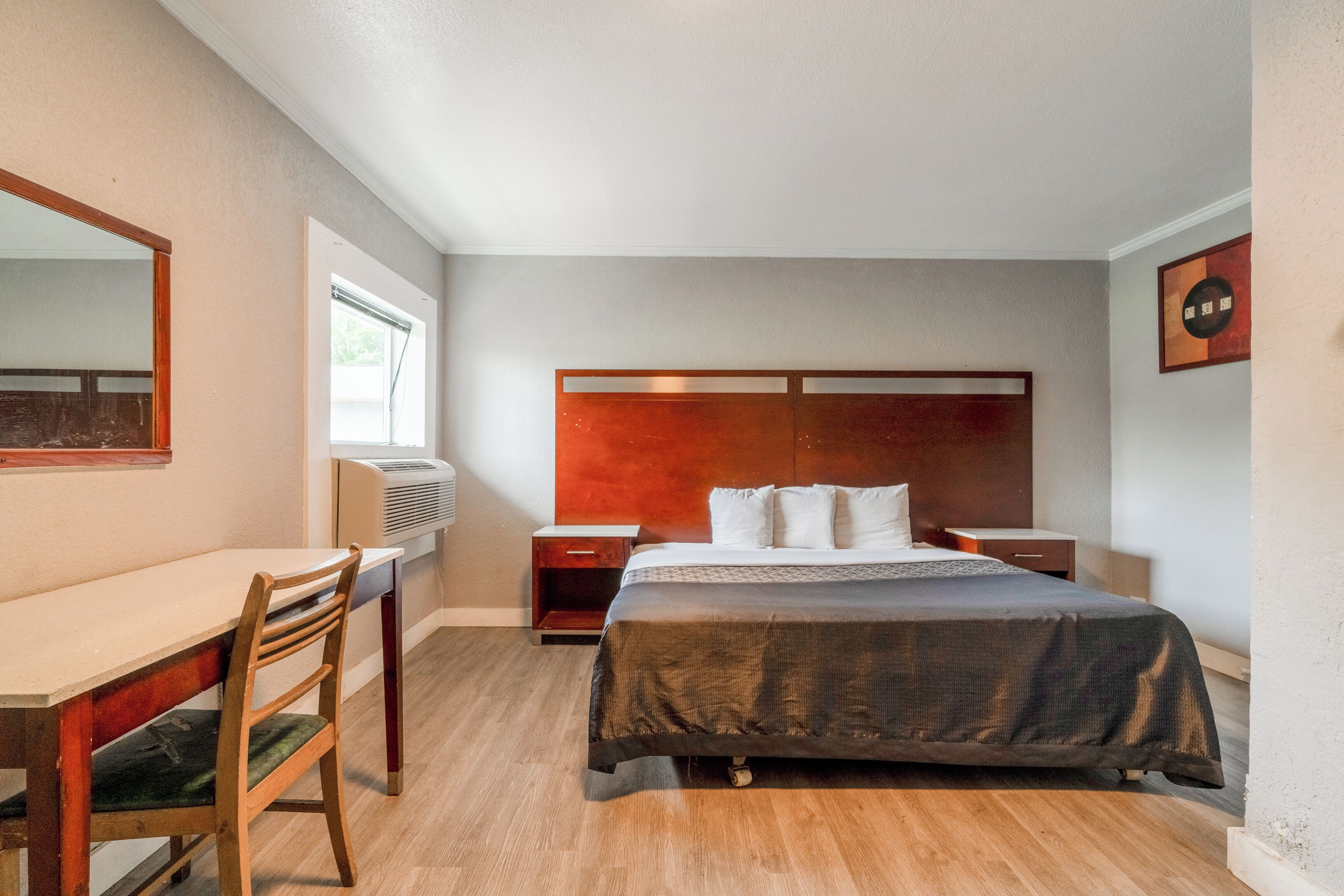 Una habitación de hotel moderna con un escritorio de madera, una cama con colcha oscura, una mesita de noche y un espejo montado en la pared.