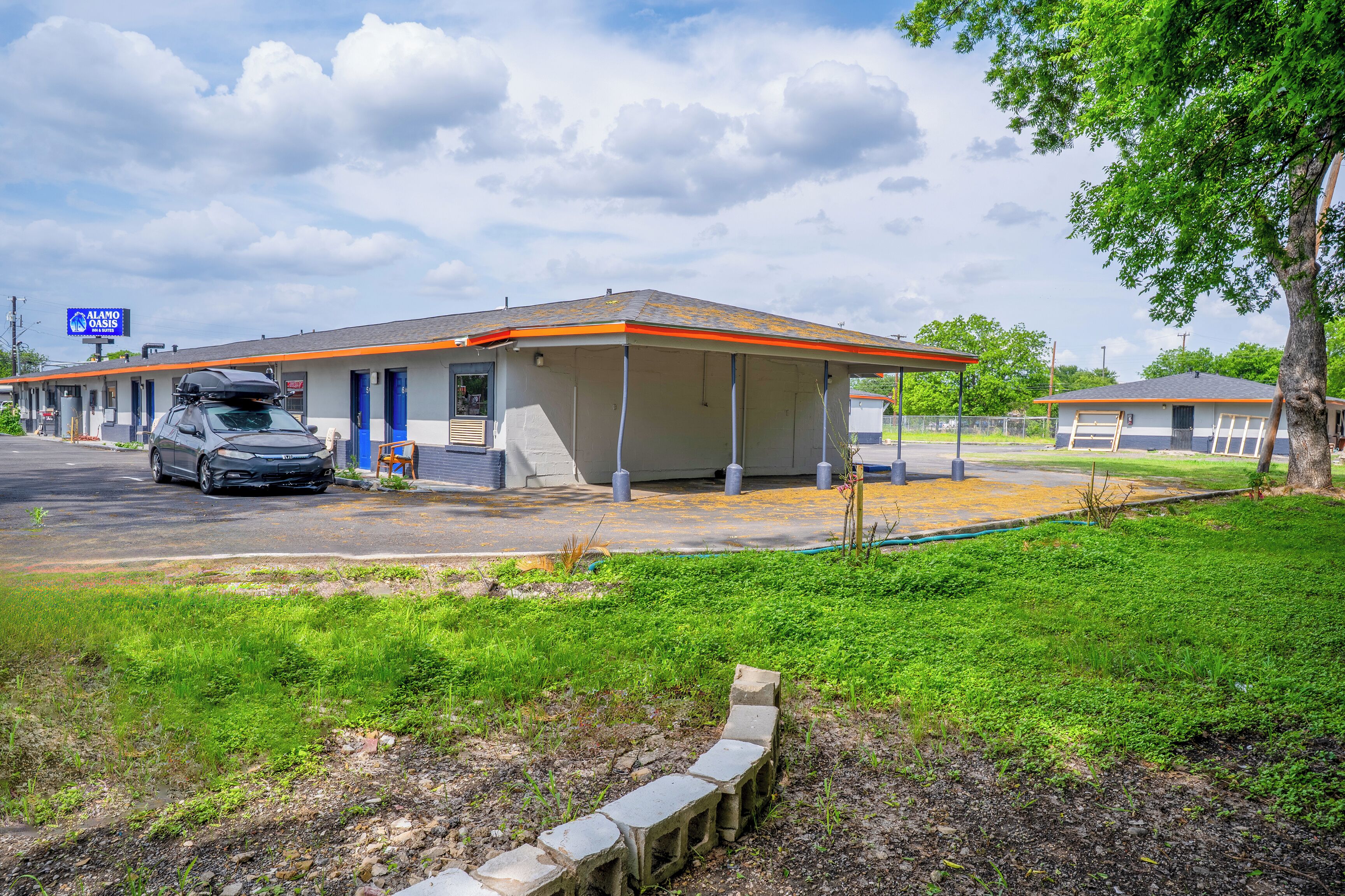 A motel with a covered parking area, a car parked outside, and a grassy area with a stone border.