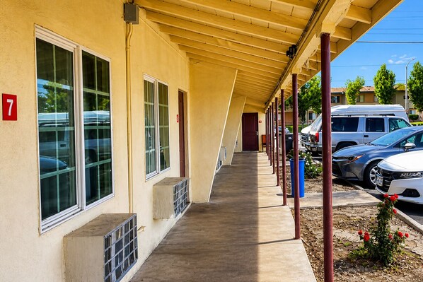 A covered outdoor corridor with a concrete walkway, a building with numbered windows, and parked cars.