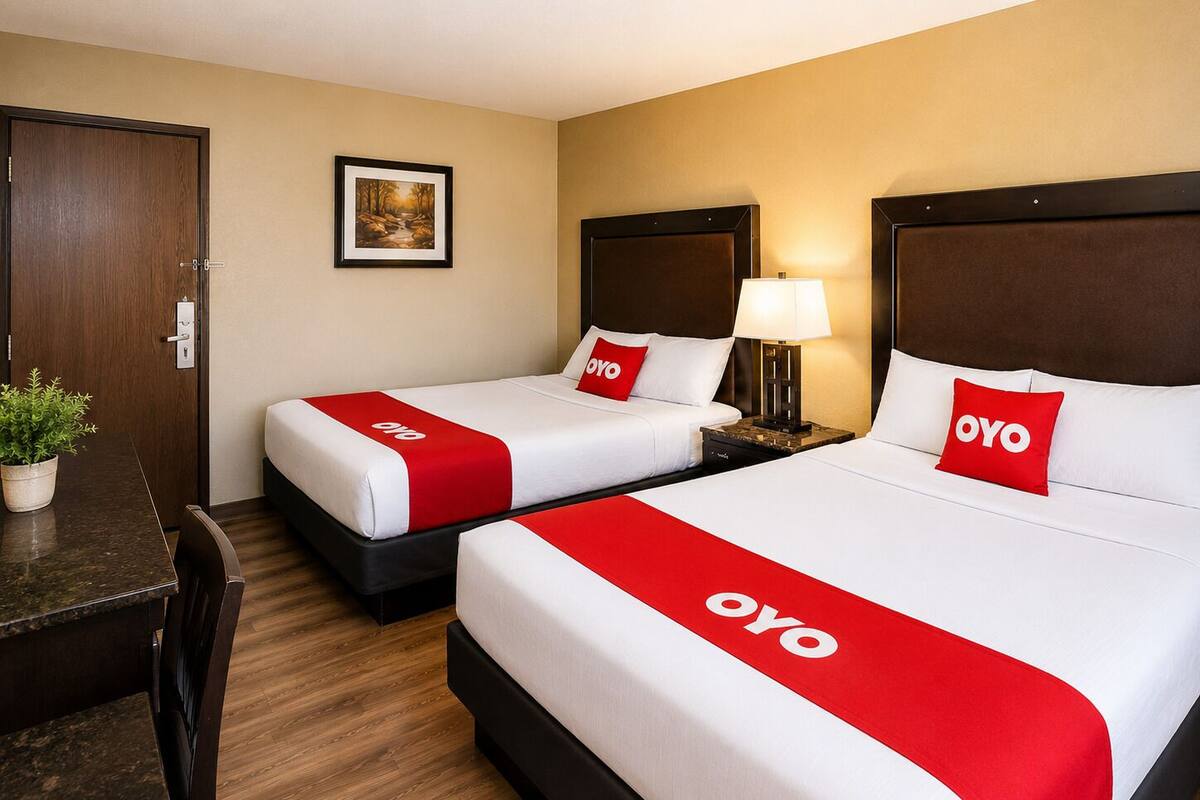 A hotel room with two beds, each with a red and white bedspread and pillows, a nightstand with a lamp, a wooden desk, and a framed picture on the wall.