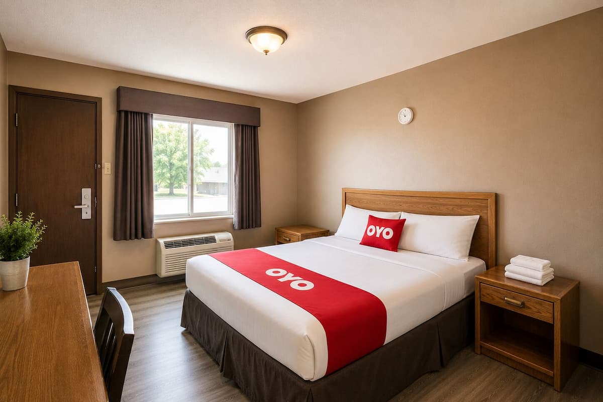 A hotel room with a bed, bedside tables, a desk, a chair, a potted plant, a window with curtains, and a wall clock.