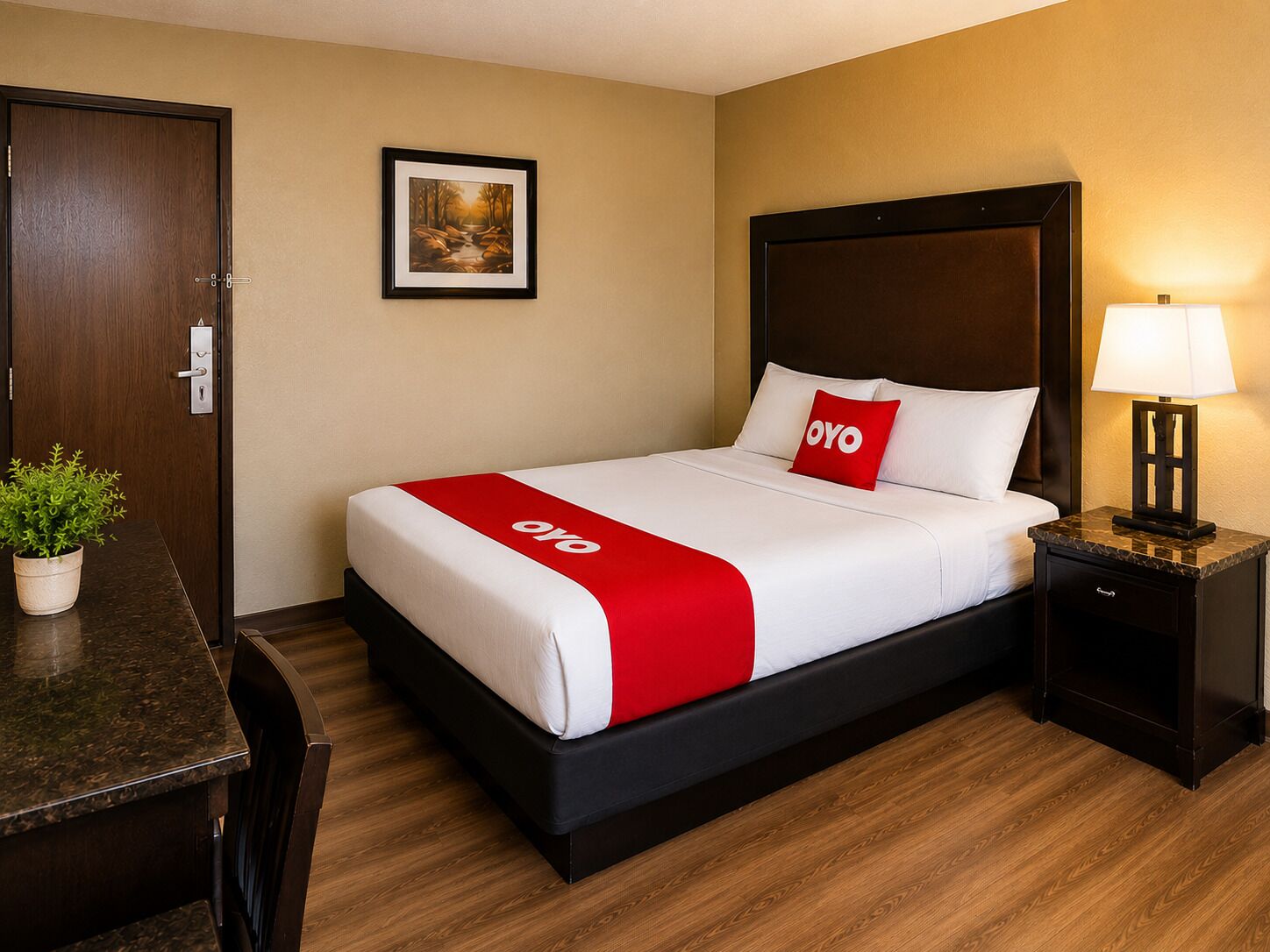 A hotel room with a bed, a nightstand, a desk, a chair, a potted plant, and a framed picture on the wall.