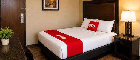 A hotel room with a bed, a nightstand, a desk, a chair, a potted plant, and a framed picture on the wall.