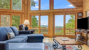 A spacious living room with a large sectional sofa, a coffee table, and a television. The room features large windows offering a view of the outdoors.