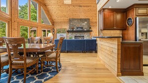 A spacious dining area with a wooden table and chairs, a stone fireplace, and a kitchen with wooden cabinets and a stainless steel refrigerator.