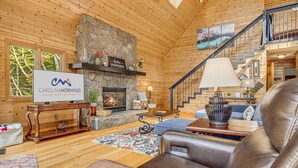 A cozy log cabin interior with a stone fireplace, a sign for Carolinamornings, and a staircase leading to an upper level.