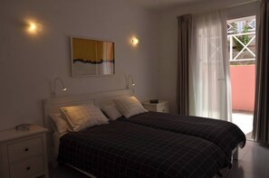 A double bed with a checkered bedspread, a bedside table with a lamp, a wall-mounted artwork, and a window with curtains. - Apt. El Guirre Nº 8 - bajo (Valle Gran Rey)