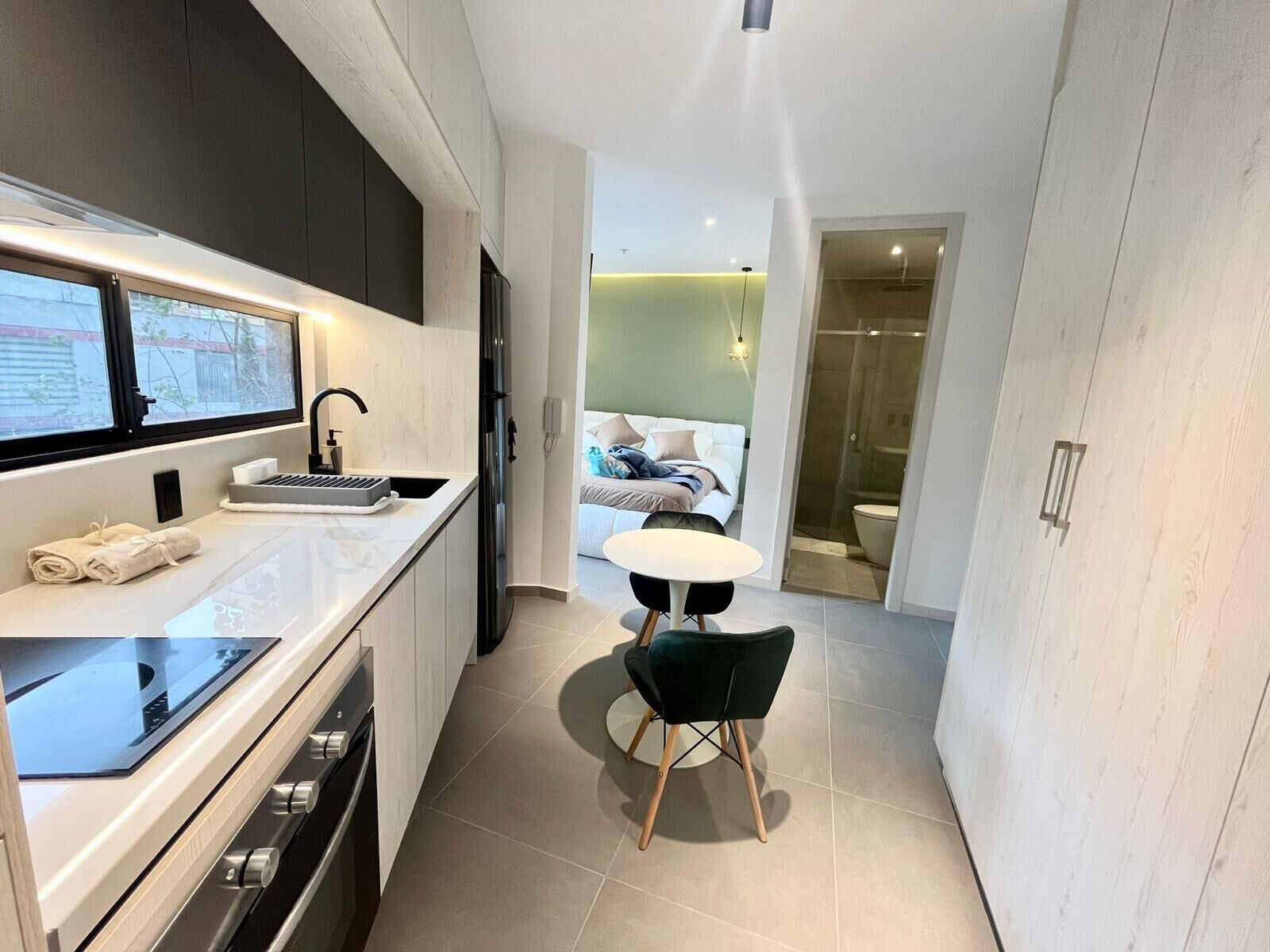 A modern hotel room with a kitchenette, a dining area, and a bedroom with a large window.