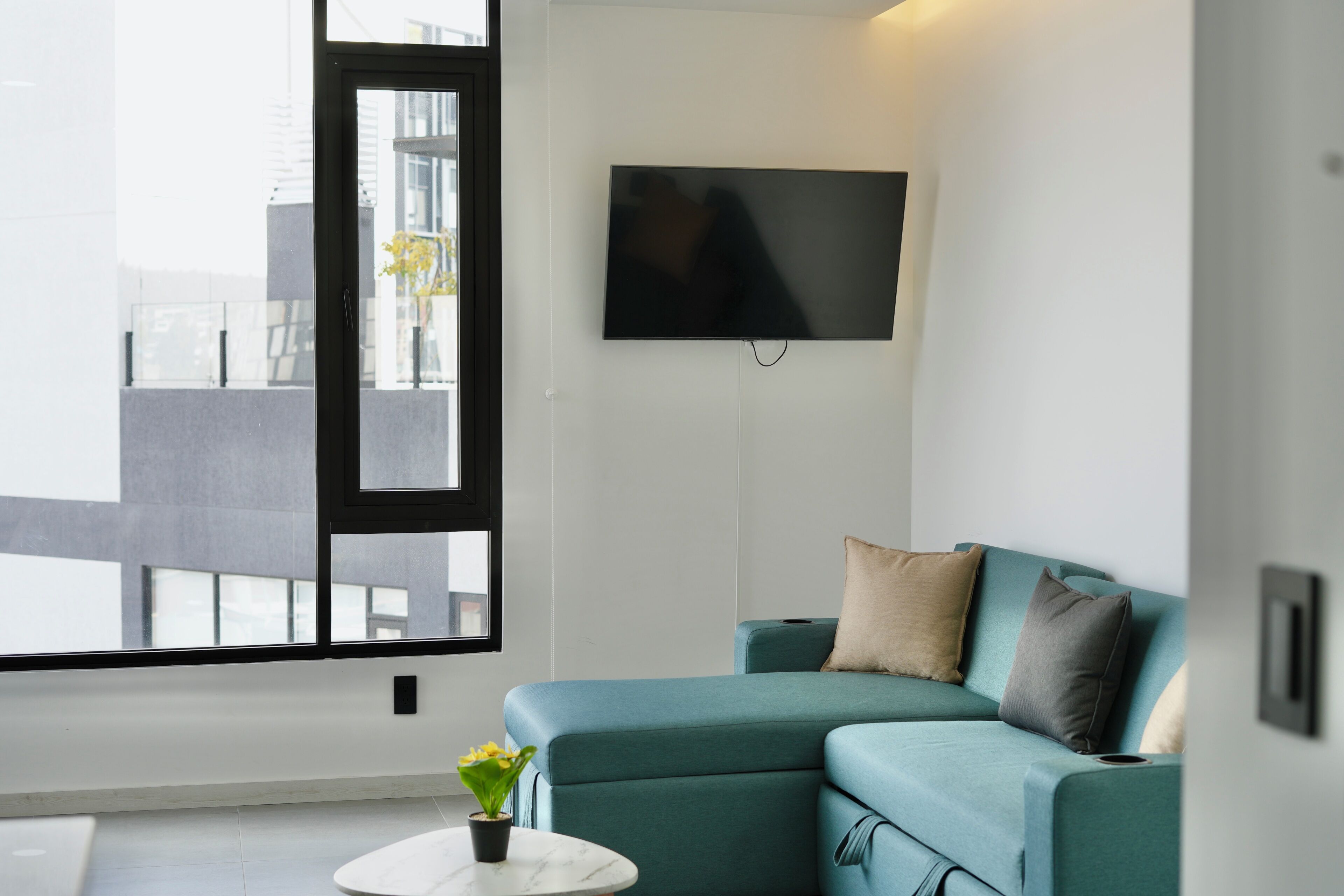Ein modernes Wohnzimmer mit einem türkisblauen Sofa, einem flachbildfernseher an der Wand, einem kleinen runden Tisch mit einem Blumentopf und einem großen Fenster mit Blick auf Gebäude.