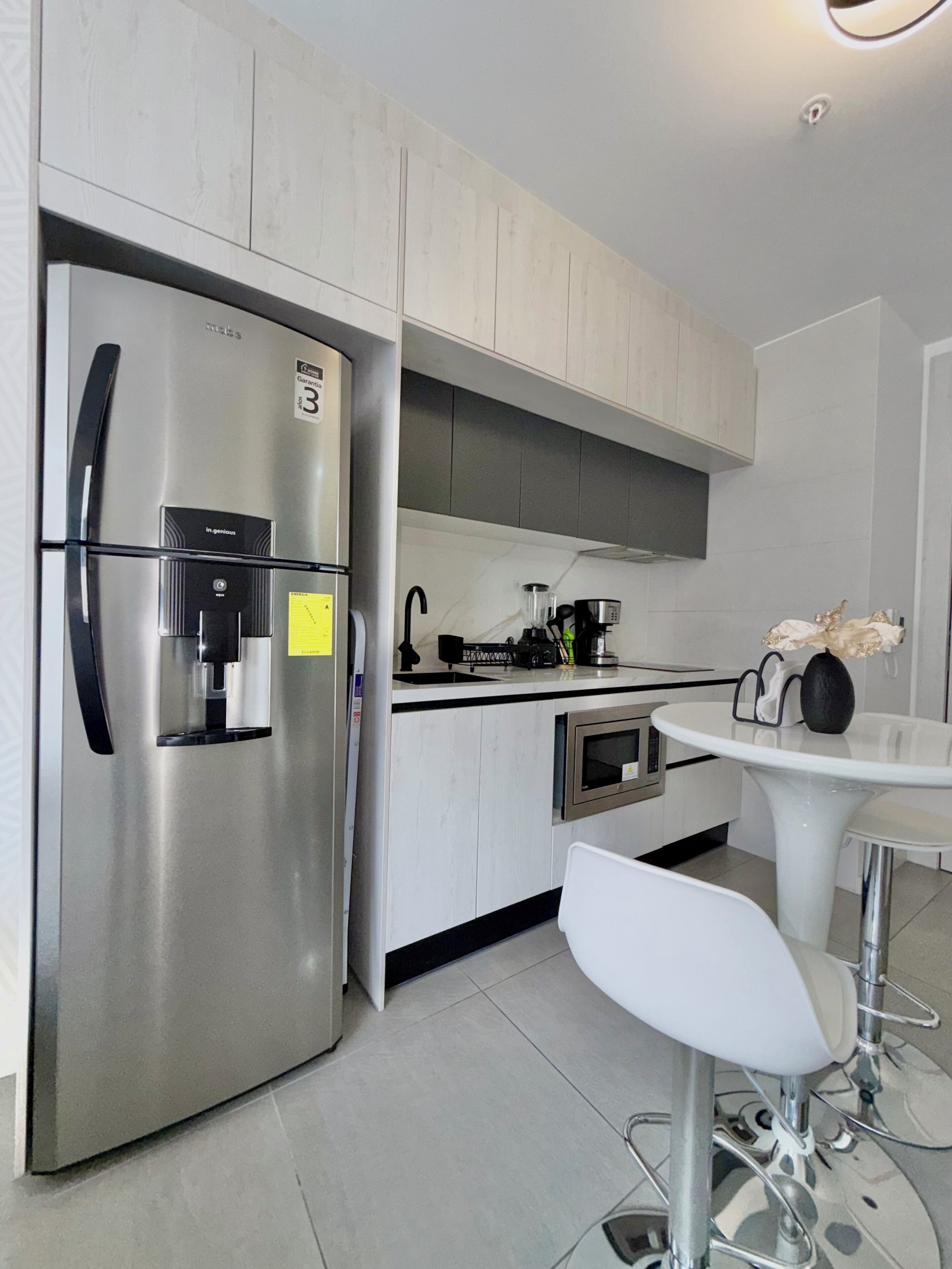 Una cocina moderna con un refrigerador de acero inoxidable, un horno empotrado, una cafetera y una mesa de comedor blanca con una silla.