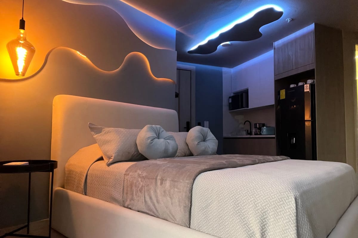Un dormitorio moderno con cama, almohadas, mesita de noche, una zona de cocina integrada y un diseño de techo original.
