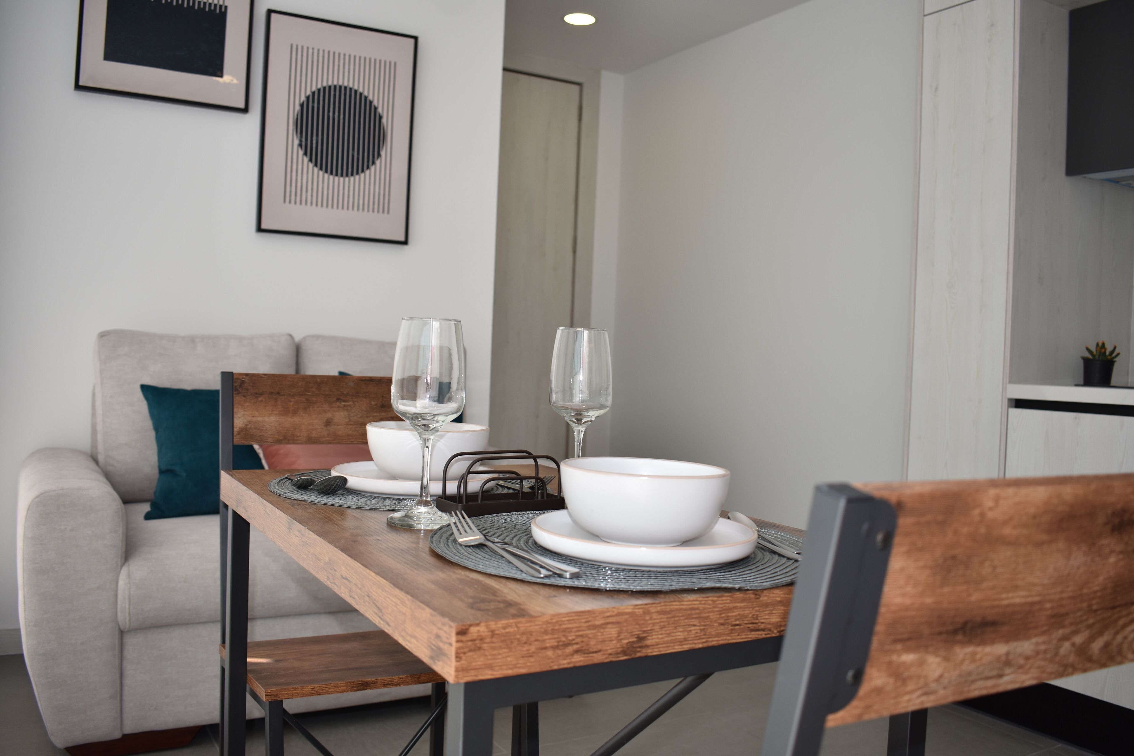 Un coin repas avec une table en bois dressée pour deux personnes, un canapé et des œuvres d’art encadrées accrochées au mur.