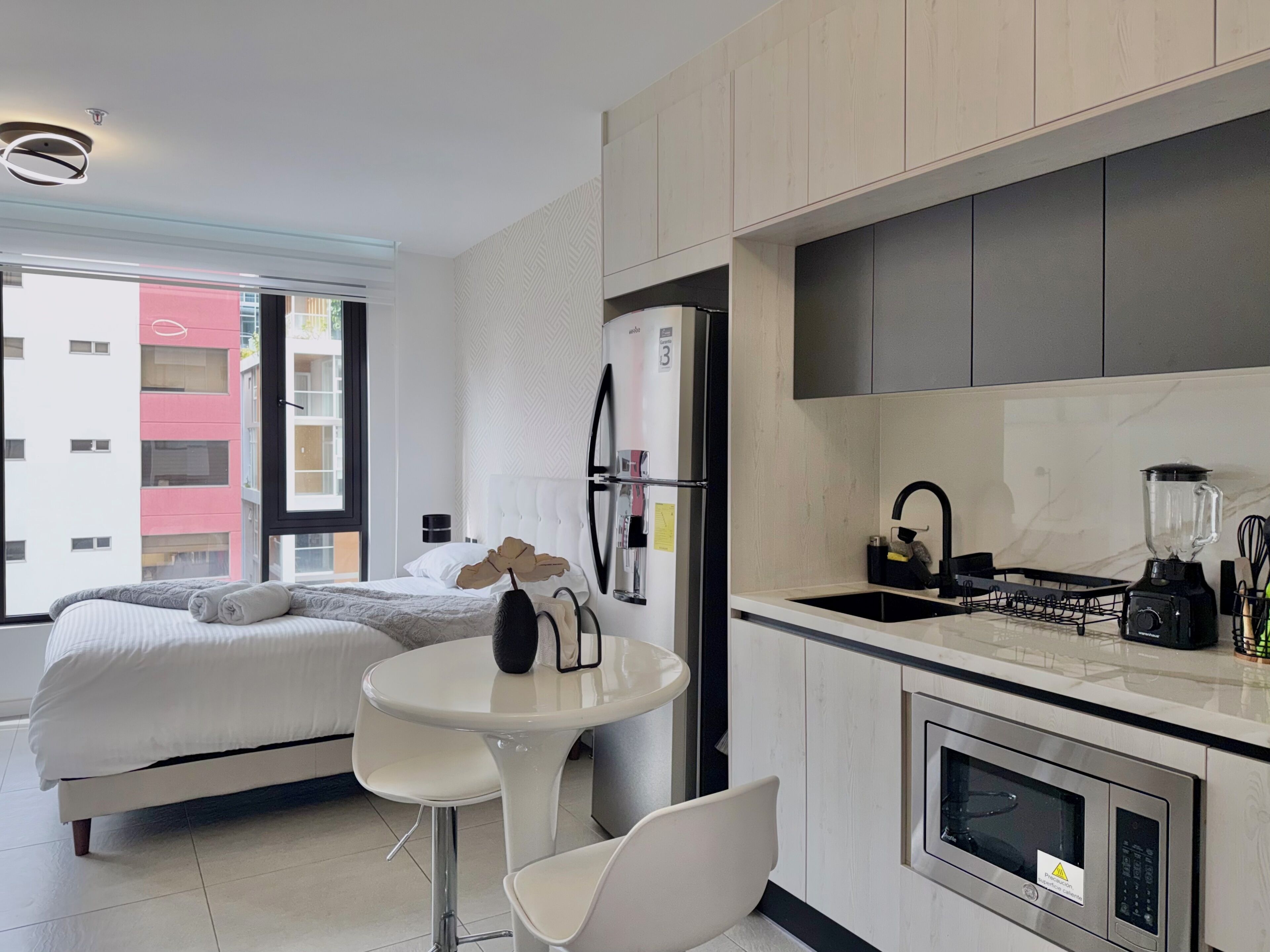 A modern hotel room with a bed, a small dining table, a kitchen area with a refrigerator, a stove, and a microwave.