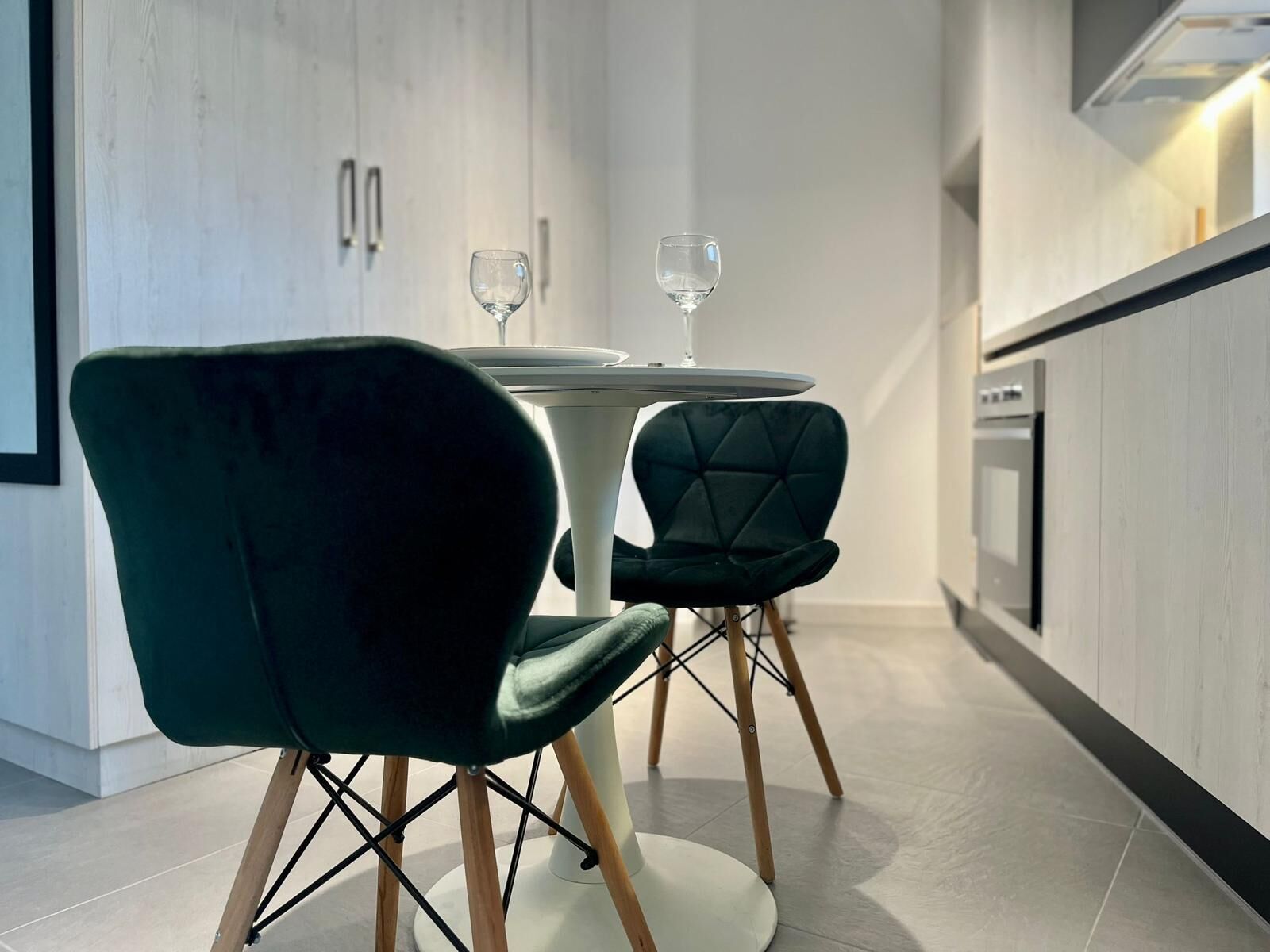 Una cocina moderna con una mesa redonda para comer, dos sillas tapizadas en verde oscuro y una copa de vino sobre la mesa.