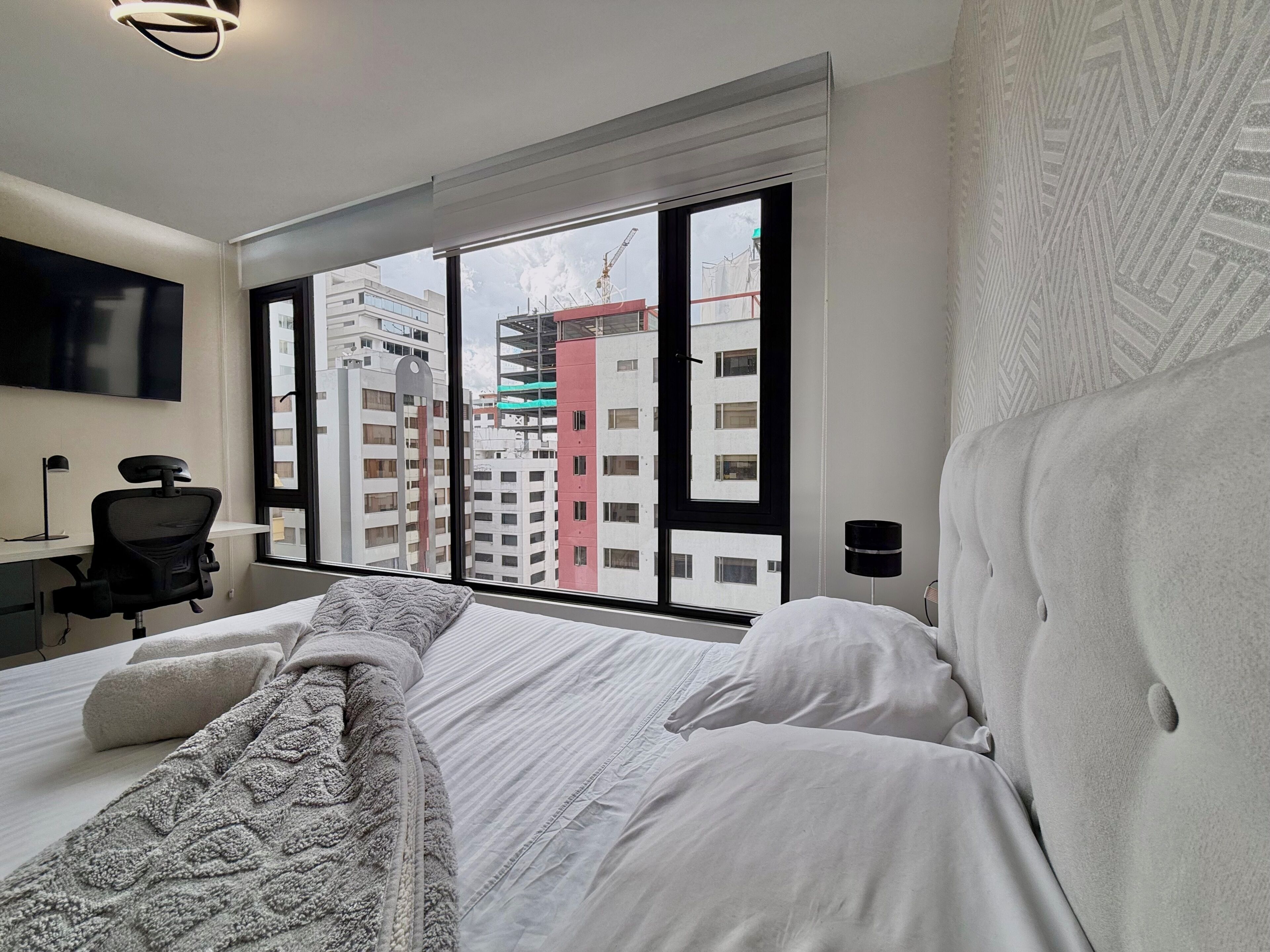 A hotel room with a large window, a bed with white linens, a desk with a chair, and a TV mounted on the wall.