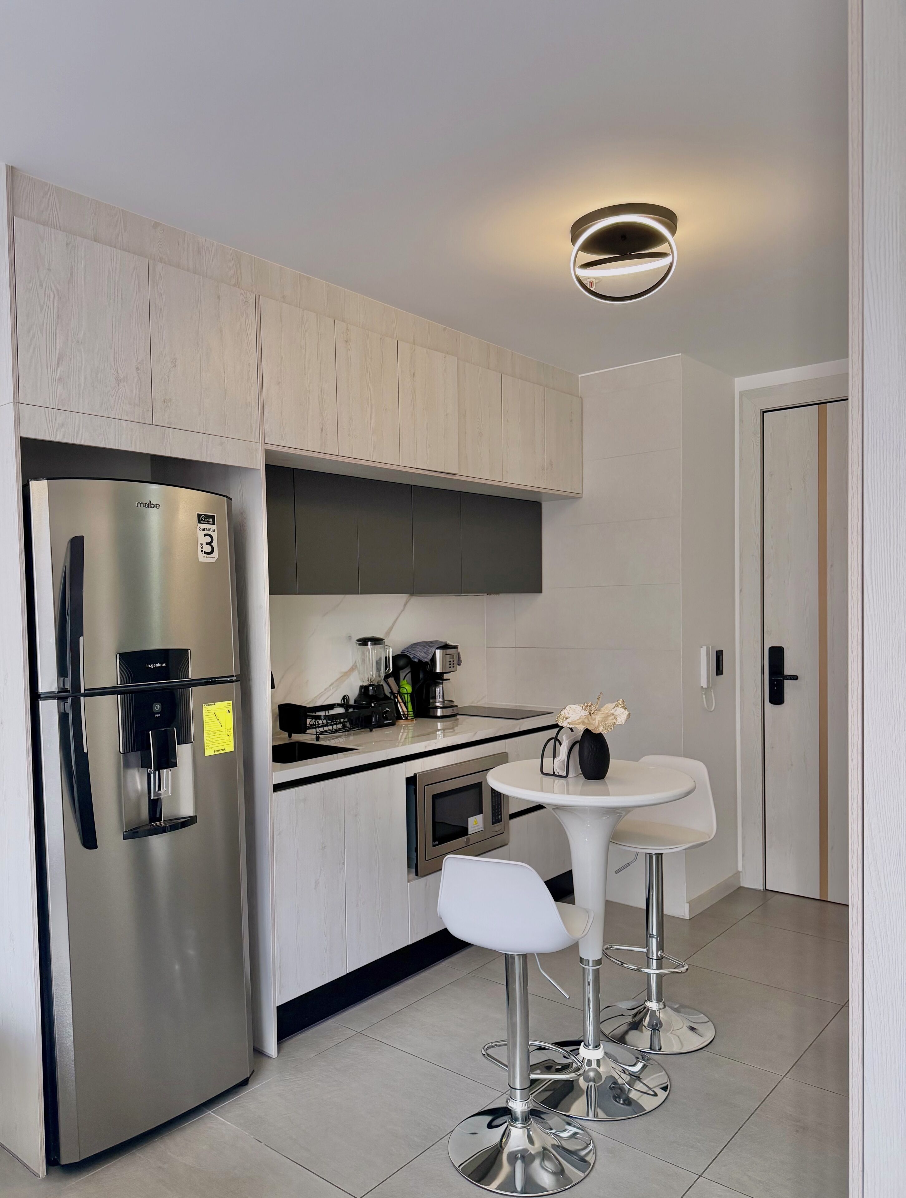 Una cocina moderna con un refrigerador de acero inoxidable, un microondas integrado, una cafetera y un dispensador de agua. Hay una mesa redonda pequeña con dos taburetes blancos.